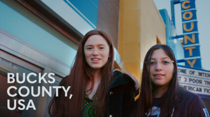 Two young girls wearing dark jackets stand in front of a movie theatre. Copy to the left reads BUCKS COUNTY, USA