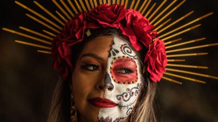 A Latina with half of her face painted in white red and black. She has a crown of roses on her head.
