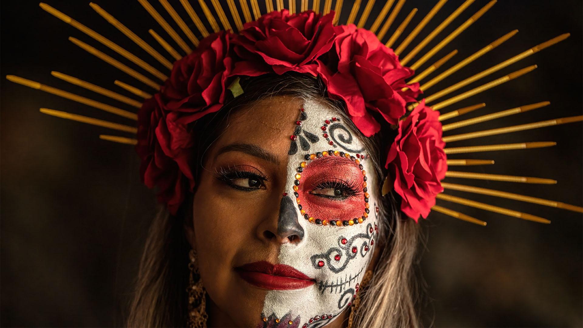 A Latina with half of her face painted in white red and black. She has a crown of roses on her head.
