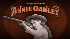 Annie Oakley wearing a hat and dress aiming a gun in front of her. About her image reads Annie Oakley in red.