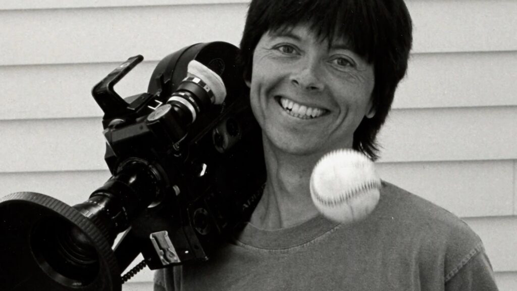 A black and white photo of Ken Burns with a camera resting on his shoulder and a baseball in the forefront