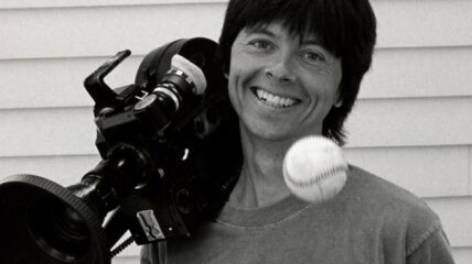 A black and white photo of Ken Burns with a camera resting on his shoulder and a baseball in the forefront