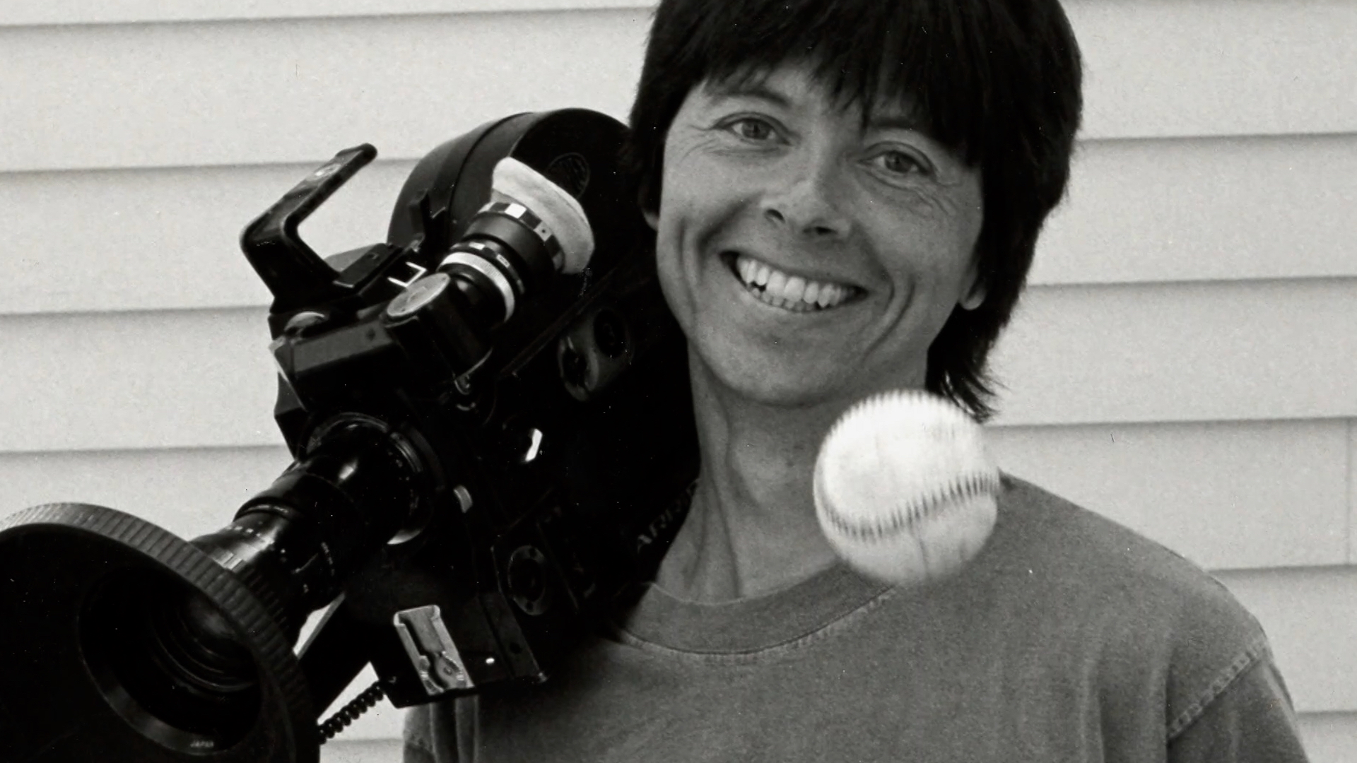A black and white photo of Ken Burns with a camera resting on his shoulder and a baseball in the forefront